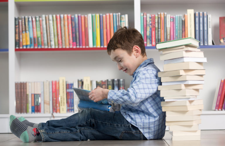 Young boy reading a stack of books in front of a bookcase