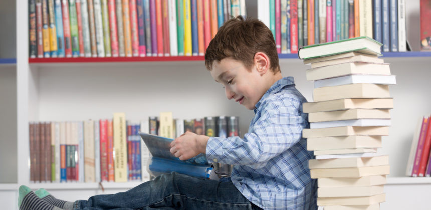 Young boy reading a stack of books in front of a bookcase