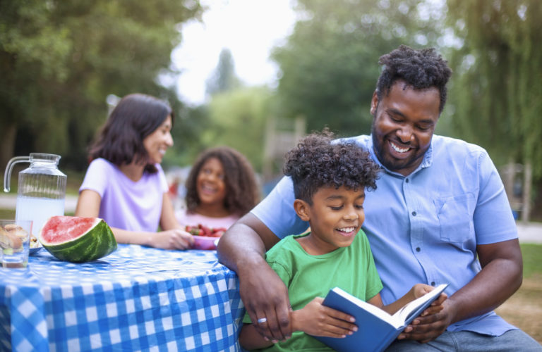 Son reading book with his African-American father at family outdoor picnic