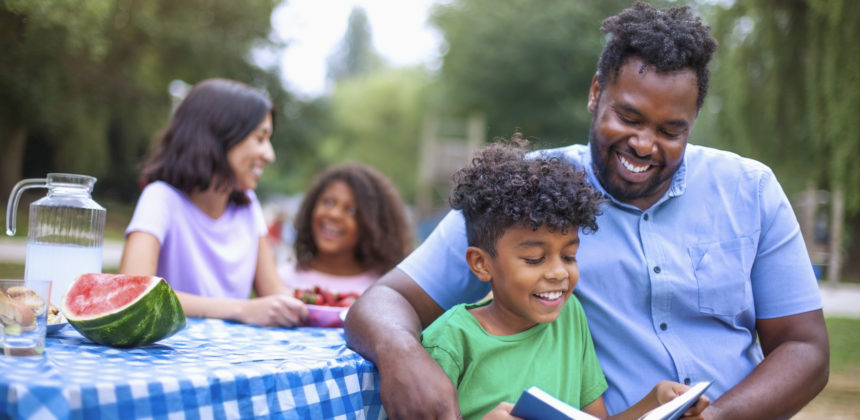 Son reading book with his African-American father at family outdoor picnic