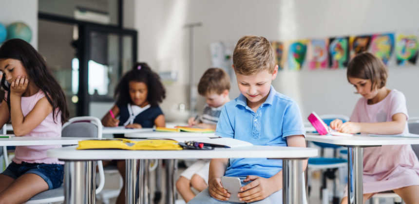 A small school boy with smartphone sitting at the desk in classroom, playing.